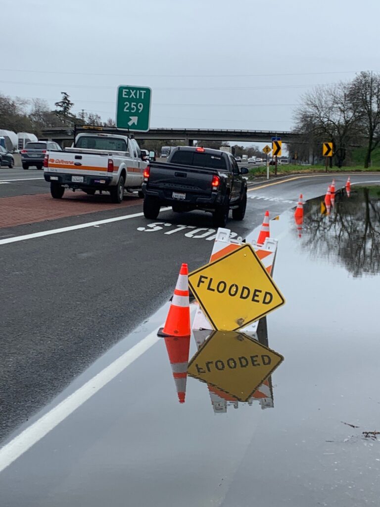 Caltrans photograph showing flooded roadway, warning sign and traffic cones alongside moving traffic